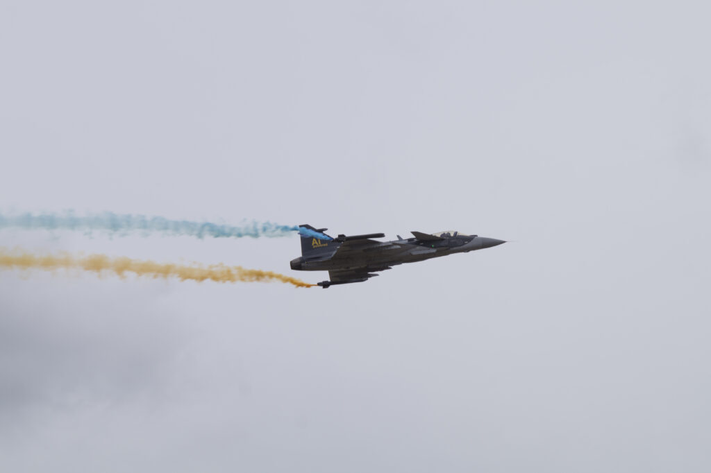 Un Saab Gripen E en vol, laissant derrière lui des traînées de fumée bleue et jaune