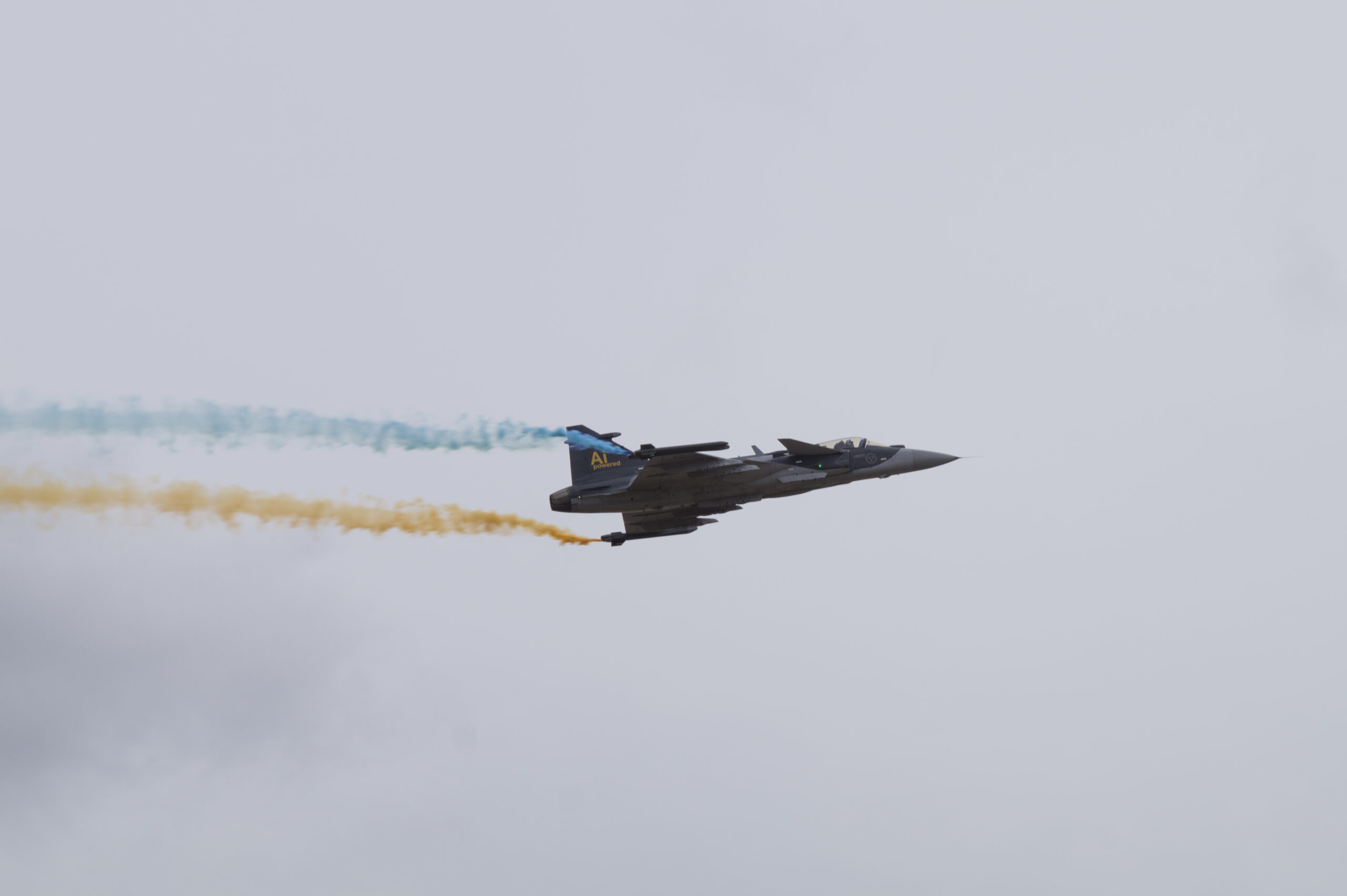 Un Saab Gripen E en vol, laissant derrière lui des traînées de fumée bleue et jaune